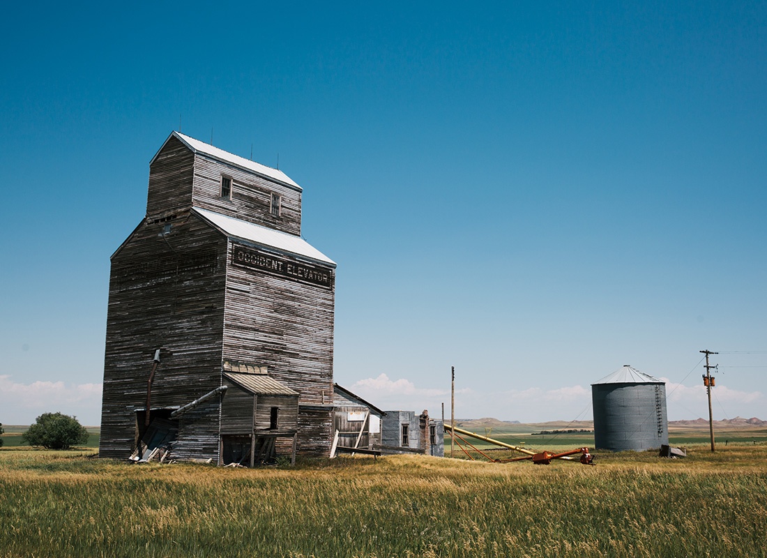 About Our Agency - View of a Grain Storage Building and Silo on a Farm in North Dakota Against a Bright Blue Sky Surrounded by a Field of Grass