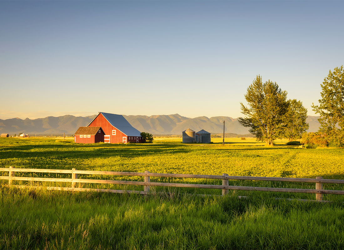 We Are Independent - Scenic Landscape of a Red Barn with a Wooden Fence on a Farm at Sunset with the Mountains Visible in the Background